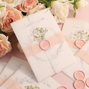 May include: Wedding invitation suite featuring white envelopes with "Save the Date" text, adorned with a pink ribbon, dried flowers, and a pink wax seal. The background includes pink roses and a soft pink surface.