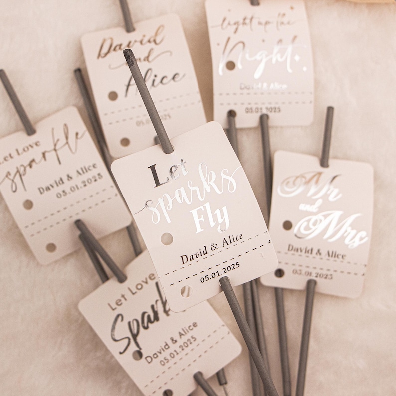 May include: A collection of sparklers with light beige tags. The tags have text in silver script, including "Let Sparks Fly," "Mr and Mrs," and names with dates. The sparklers are gray.