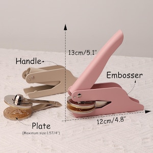 May include: A pink metal embosser with a handle and a round plate. The embosser is 13 inches long and 4.8 inches wide. The plate is 1.57 inches in diameter. The embosser is used to create raised lettering or designs on paper or other materials.