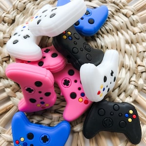 May include: A collection of colorful silicone game controller charms in pink, blue, black, and white. The charms are shaped like video game controllers with buttons and a directional pad.
