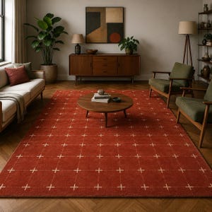 May include: A large rectangular area rug in a deep terracotta red color featuring a repeating grid pattern of thin white lines and small cross symbols. The rug is positioned on a wooden floor in a room with a sofa, armchairs, and a coffee table.