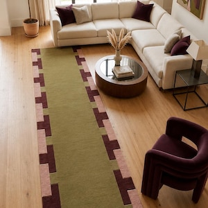 May include: A long rectangular area rug featuring a solid olive green center panel bordered by a geometric pattern of alternating dark burgundy and dusty pink squares. The rug has a low-pile texture and is positioned on a light wood floor.