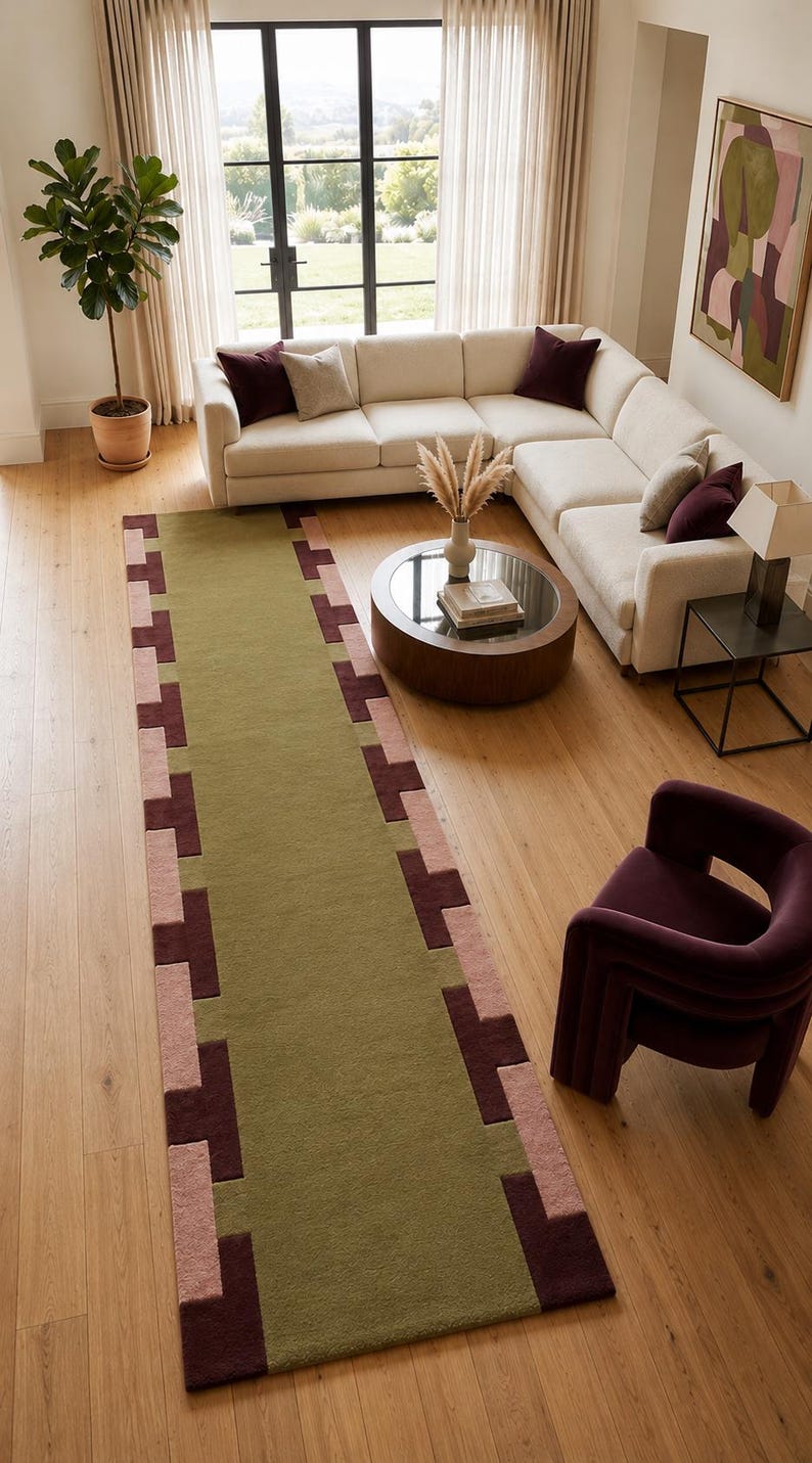 May include: A long rectangular area rug featuring a solid olive green center panel bordered by a geometric pattern of alternating dark burgundy and dusty pink squares. The rug has a low-pile texture and is positioned on a light wood floor.