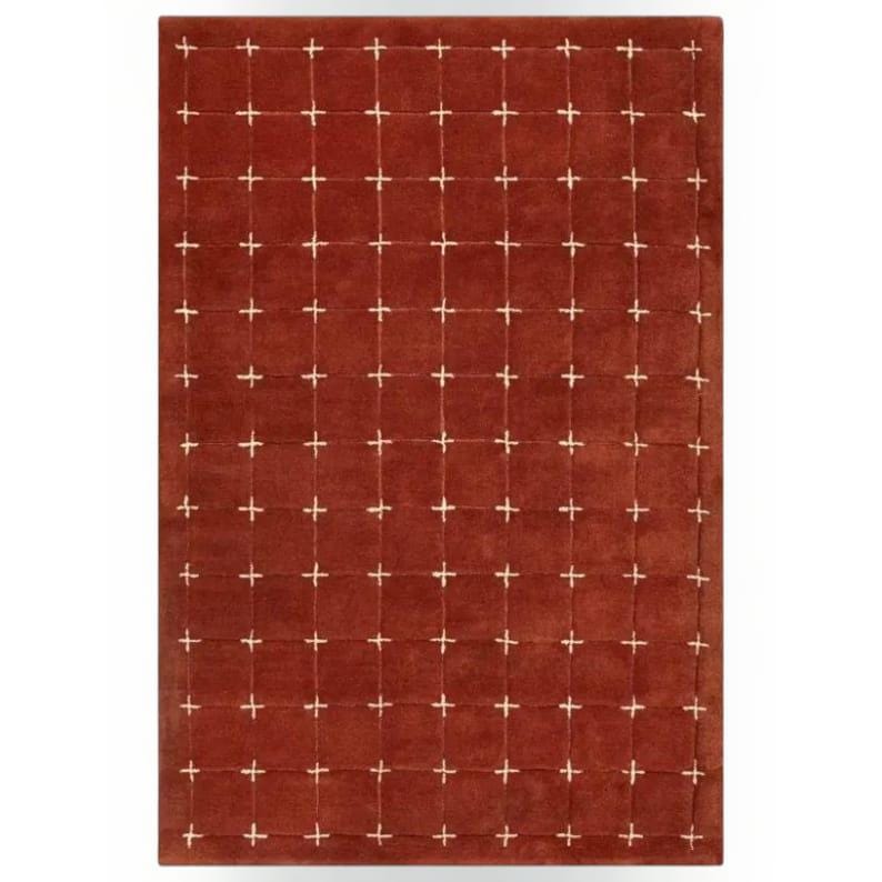 May include: A rectangular area rug in a deep terracotta red color featuring a grid pattern formed by thin, light cream-colored lines that intersect at regular intervals to create a series of uniform squares across the entire surface.