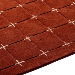 May include: A deep rust-colored area rug featuring a repeating grid pattern of raised square sections. The squares are separated by thin, recessed white lines that intersect at small cross-shaped points, creating a textured, geometric surface design.