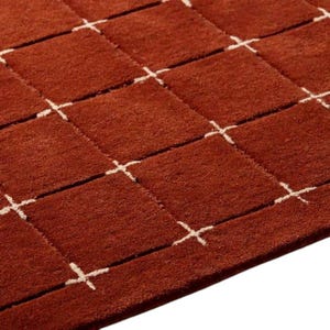 May include: A deep rust-colored area rug featuring a repeating grid pattern of raised square sections. The squares are separated by thin, recessed white lines that intersect at small cross-shaped points, creating a textured, geometric surface design.