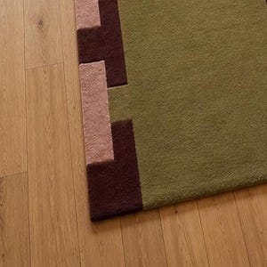 May include: A rectangular area rug featuring a large olive green central field with a textured surface. The border displays a geometric pattern composed of alternating dark brown and light pink blocks arranged in a stepped, interlocking design.