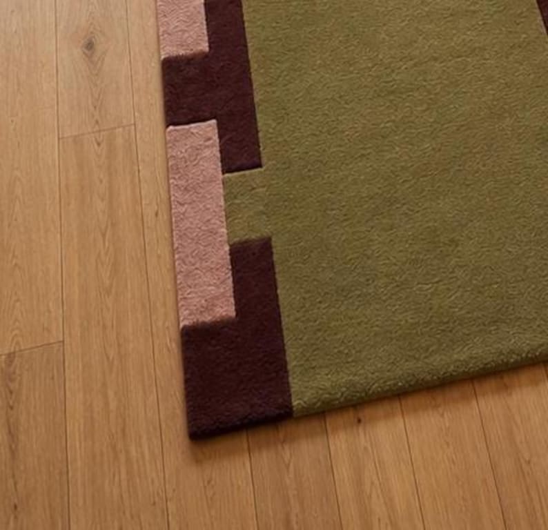 May include: A rectangular area rug featuring a large olive green central field with a textured surface. The border displays a geometric pattern composed of alternating dark brown and light pink blocks arranged in a stepped, interlocking design.