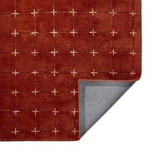 May include: A rectangular rug with a deep rust-colored surface featuring a grid pattern of thin, light-colored lines. The lines intersect at small cross-shaped marks. One corner of the rug is folded back to reveal a gray, textured backing material.