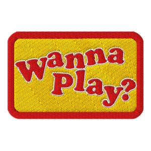 May include: Embroidered patch with the text "Wanna Play?" in red, outlined in white, on a yellow background. The patch has a red border, creating a rectangular shape. The design has a playful and inviting feel.