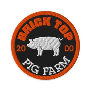 May include: Embroidered patch with a black center and an orange border. The patch features the text "BRICK TOP" in orange, and "PIG FARM" in white. A white pig is in the center, with the numbers "20" and "00" on either side.