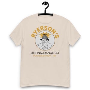 May include: A cream-coloured t-shirt with a graphic of a man in a hat and glasses inside a circle. The text reads "RYERSON'S" above and "LIFE INSURANCE CO." below, with "BING!" and "Punxsutawney - PA" also visible. The shirt hangs from a black hanger.