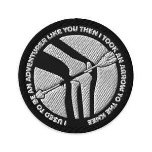 Puede incluir: Parche bordado en blanco y negro con el texto "I USED TO BE AN ADVENTURER LIKE YOU THEN I TOOK AN ARROW TO THE KNEE." El parche presenta una imagen estilizada de una rodilla con una flecha.