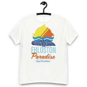 May include: White t-shirt with a blue and orange graphic of a spaceship in front of a sun and waves. The text "Fhlostorn Paradise - Legend of the Angel Constellation" is printed below the graphic.