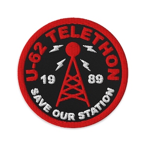 Puede incluir: Parche bordado circular con borde rojo y centro negro. El parche presenta el texto "U-62 TELETHON" en blanco, con "19" y "89" debajo. Un gráfico de torre de radio roja está en el centro, con "SAVE OUR STATION" en blanco.