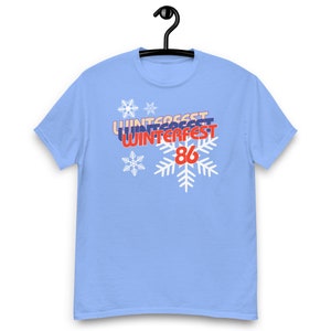 Könnte beinhalten: Hellblaues T-Shirt mit dem Text "WINTERFEST 86" in Rot, Blau und Weiß. Das Design beinhaltet Schneeflocken. Das Shirt hängt an einem schwarzen Kleiderbügel.
