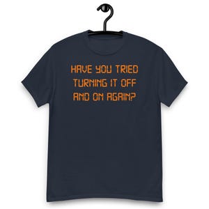 Op de afbeelding: Een donker marineblauw t-shirt met korte mouwen en de oranje tekst "HAVE YOU TRIED TURNING IT OFF AND ON AGAIN?" in een gepixeld lettertype op het midden van de borst. Het shirt hangt aan een zwarte plastic kledinghanger.