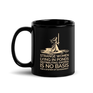 Könnte beinhalten: Schwarze Keramik-Tasse mit einer weißen Grafik einer bekleideten Figur, die ein Schwert hält, mit dem Text "STRANGE WOMEN LYING IN PONDS DISTRIBUTING SWORDS IS NO BASIS FOR A SYSTEM OF GOVERNMENT."