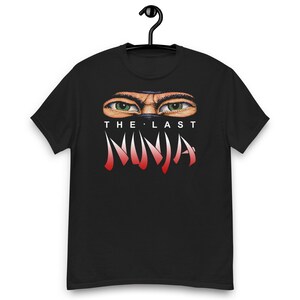 Peut inclure: Un t-shirt noir à manches courtes orné d'un graphisme représentant deux yeux verts regardant derrière un masque sombre. Sous les yeux, le texte THE LAST NINJA est inscrit en lettres stylisées blanches et rouges au centre de la poitrine.