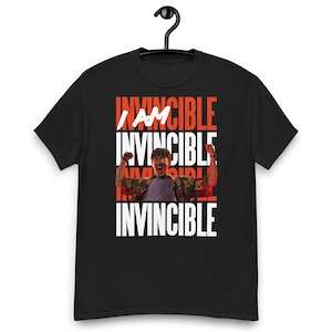 Pode incluir: Camiseta preta com as palavras "I AM INVINCIBLE" em letras brancas e vermelhas. A camiseta apresenta uma foto de uma pessoa com os braços levantados em uma pose de celebração. A camiseta está pendurada em um cabide preto.
