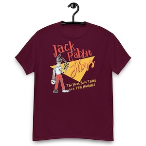 May include: A maroon short-sleeve t-shirt featuring a graphic of a cartoon rabbit holding a trophy. The text reads Jack Rabbit Slim's and The Next Best Thing to a Time Machine. The design includes a yellow triangle and red and white accents.