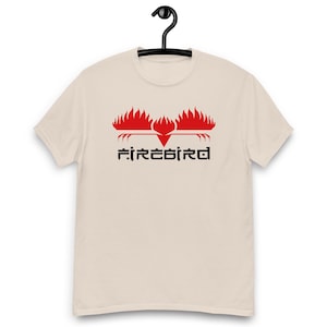 May include: A beige short-sleeved t-shirt featuring a graphic of a stylised red bird made of flames above the black text "FIREBIRD" in a geometric font. The design is centred on the chest of the garment, which is displayed on a black hanger.