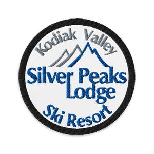Pode incluir: Remendo bordado circular com o texto "Kodiak Valley Silver Peaks Lodge Ski Resort" em azul e cinza sobre um fundo branco. O remendo tem uma borda preta e um gráfico de montanha.