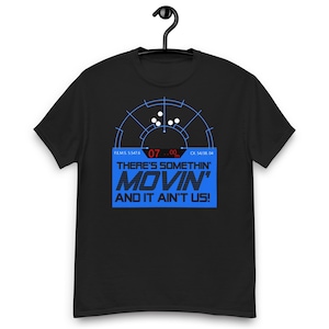 May include: Black t-shirt with a blue graphic design. The design includes a radar-like image with the text "THERE'S SOMETHIN' MOVIN' AND IT AIN'T US!" in blue. The shirt is hanging on a black hanger.
