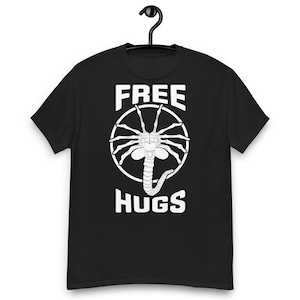 Könnte beinhalten: Ein schwarzes Kurzarm-T-Shirt mit einer weißen Grafik eines Facehugger-Wesens, das in einem Kreis zentriert ist. Der Text "FREE" ist über der Grafik und "HUGS" unter der Grafik in einer fetten, weißen, serifenlosen Schriftart aufgedruckt.