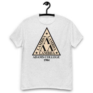 Peut inclure: Un t-shirt gris clair avec un logo triangulaire noir "Lambda", le texte "LAMBDA" et "ADAMS COLLEGE 1984". Le t-shirt est suspendu à un cintre noir.