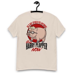 Op de afbeelding: Een crèmekleurig t-shirt met een afbeelding van een varken met een ronde bril en een bliksemschicht-litteken op het voorhoofd. De tekst luidt: HE'S NOT SPIDER-PIG ANYMORE HES HARRY PLOPPER NOW, tegen een rode cirkelvormige achtergrond.