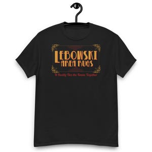 May include: A black short-sleeved t-shirt featuring a graphic print with the text "LEBOWSKI AREA RUGS" in orange and yellow, enclosed in a decorative border, with the red text "It Really Ties the Room Together" printed underneath the design.
