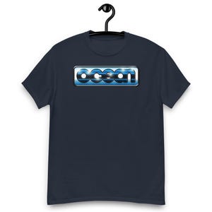 Op de afbeelding: Een donkerblauw t-shirt met korte mouwen en een rechthoekige afbeelding op de borst. De afbeelding toont het woord ocean in een gestileerd, metallic blauw lettertype met een glanzende, reflecterende afwerking in een zilverkleurig kader.