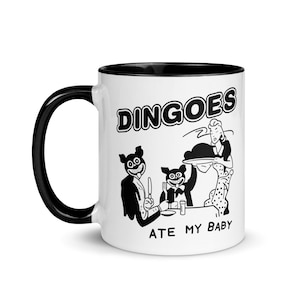 Könnte beinhalten: Weißer Keramikbecher mit schwarzem Henkel und Innenraum. Der Becher zeigt eine Schwarz-Weiß-Cartoon-Illustration von anthropomorphen Dingos an einem Tisch mit dem Text "DINGOES ATE MY BABY".