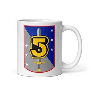 Puede incluir: Una taza de cerámica blanca con un emblema en forma de escudo dividido diagonalmente en secciones gris y azul. El emblema muestra un gran número 5 amarillo, una espada plateada vertical y cinco estrellas blancas, bordeado por una línea roja.