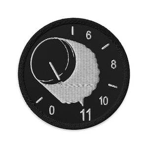 Puede incluir: Parche bordado en blanco y negro con un diseño de dial. El dial muestra los números 0, 6, 8, 10 y 11, con una aguja blanca que indica una posición. El parche tiene un borde negro.