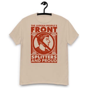 May include: Beige short-sleeve t-shirt with a red graphic. The graphic features the text "THE JUDEAN PEOPLE'S FRONT" above a circular image of a person with a laurel wreath, crossed out by a line. Below the image is the text "SPLITTERS AND PROUD".