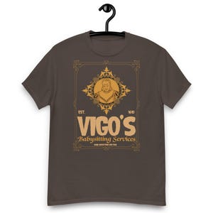 May include: A brown t-shirt with a gold graphic featuring a regal figure and the text "VIGO'S Babysitting Services." The design includes the text "EST. 1610" and "HOME ABDUCTIONS ARE FREE."