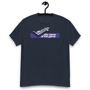 May include: A navy blue short-sleeve t-shirt featuring a graphic print on the chest. The design includes the word Imagine in a metallic blue font above a purple horizontal bar containing the white text the name of the game.