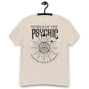 May include: Beige t-shirt with a black graphic featuring the text "WORLD OF THE PSYCHIC" above an illustration of a man, an eye within a triangle, and the words "WITH DR. PETER VENKMAN". The shirt is hanging on a black hanger.