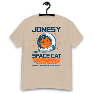 May include: A beige short-sleeved t-shirt with a graphic of an orange cat in a space helmet. The text above the cat reads "JONESY", and below it, "THE SPACE CAT". Below the cat graphic is the text "THE LAST SURVIVOR OF THE NOSTROMO".
