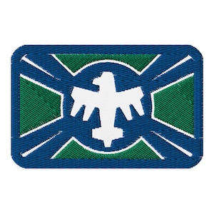 Może przedstawiać: Naszywka haftowana w kolorze niebieskim i zielonym z białym orłem w środku. Orzeł otoczony jest zielonym okręgiem i niebieskim krzyżem.