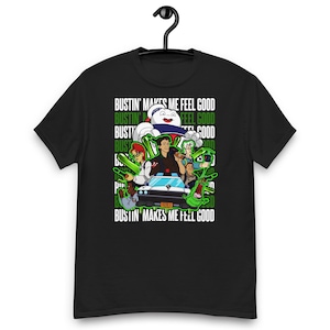 May include: A black short-sleeved t-shirt featuring a graphic print of Ghostbusters characters, the Ecto-1 vehicle, and the Stay Puft Marshmallow Man. The text "BUSTIN' MAKES ME FEEL GOOD" is repeated in white and green against a dark background.