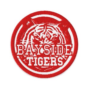 May include: A round, red embroidered patch with a white tiger illustration and the words "BAYSIDE TIGERS" in white block letters. The patch has a white border and is likely for apparel.
