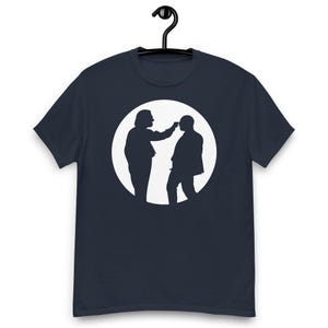 May include: Navy blue t-shirt featuring a white graphic of two people in silhouette, one touching the other's forehead, set within a white circle. The t-shirt is hanging on a black hanger.