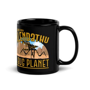 May include: A black ceramic mug featuring a graphic print with the text GREETINGS FROM KLENDATHU BUG PLANET in yellow. The illustration depicts a yellow and black striped insectoid creature standing on a desert landscape with brown rock formations.