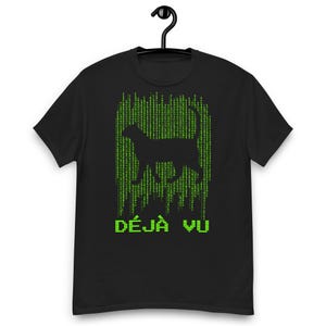 Puede incluir: Camiseta negra con la silueta de un gato y un diseño de lluvia digital verde. El texto francés "DÉJÀ VU" está impreso en verde debajo del gráfico del gato. La camiseta está colgada en una percha negra.