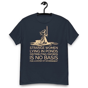 May include: A navy blue t-shirt featuring a graphic of a woman holding a sword standing in a pond. The text on the shirt reads "Strange women lying in ponds distributing swords is no basis for a system of government."