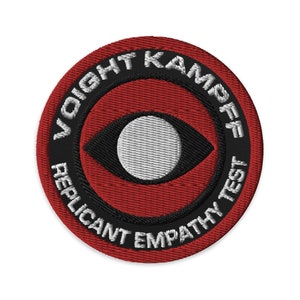 Puede incluir: Un parche circular rojo, negro y blanco con el texto "Voight-Kampff Replicant Empathy Test" alrededor del borde. El centro del parche presenta un ojo estilizado con un iris negro y una pupila blanca.