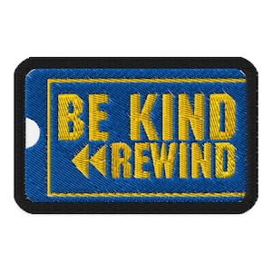 May include: Embroidered patch with the words "BE KIND REWIND" in yellow thread on a blue background. The patch has a black border and a white dot on the left side. The text is in a bold, retro style.